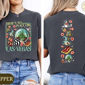 May include: Dark gray t-shirt with a colorful graphic featuring the text "THERE'S NO PLACE LIKE LAS VEGAS." The design includes a crystal ball, ruby slippers, and other Wizard of Oz-themed elements. The back of the shirt has a vertical design with similar graphics.