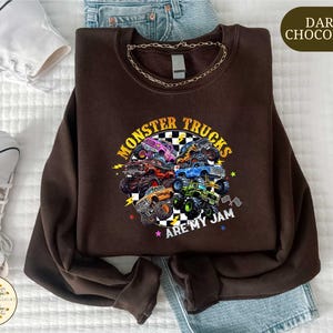 a brown sweatshirt with a graphic design of monster trucks and the text "MONSTER TRUCKS ARE MY JAM" printed on it. The sweatshirt is laid out on a bed, along with a pair of jeans and a pair of sneakers.