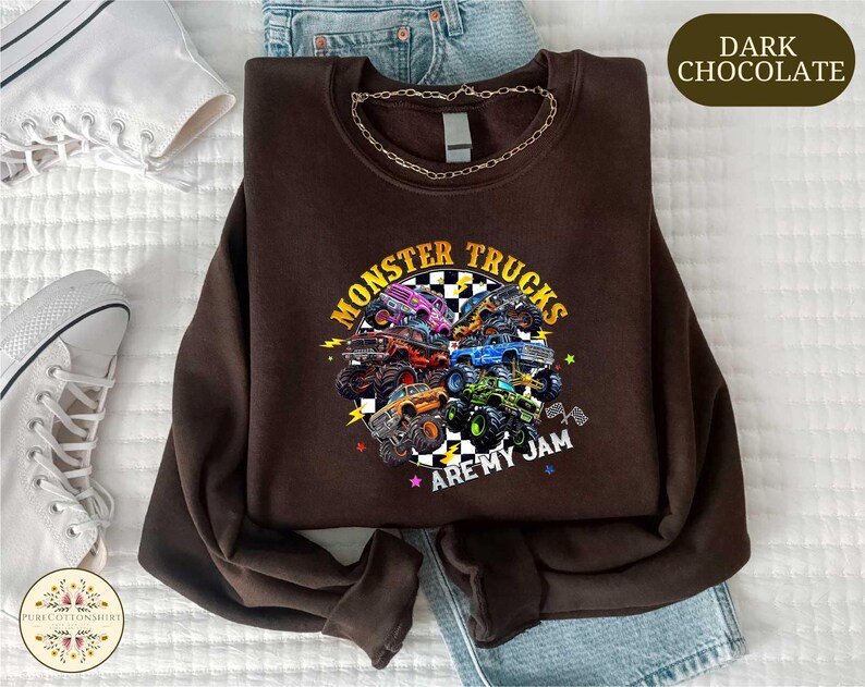a brown sweatshirt with a graphic design of monster trucks and the text "MONSTER TRUCKS ARE MY JAM" printed on it. The sweatshirt is laid out on a bed, along with a pair of jeans and a pair of sneakers.