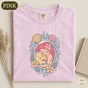 A pink t-shirt with a colorful illustration of a mermaid and a fish, surrounded by flowers and a frame.