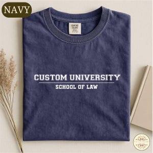 Könnte beinhalten: Marineblaues T-Shirt mit dem weißen Text "CUSTOM UNIVERSITY SCHOOL OF LAW". Das Shirt hat einen Rundhalsausschnitt und ein Etikett mit der Aufschrift "COMFORT COLORS". Das Wort "NAVY" befindet sich in einem goldenen Oval oben links.