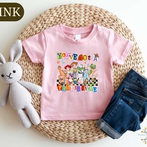 A pink t-shirt with a colorful graphic of characters from Toy Story, along with a crocheted stuffed rabbit and a pair of blue jeans, all placed on a woven placemat.