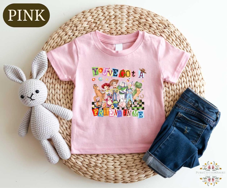 A pink t-shirt with a colorful graphic of characters from Toy Story, along with a crocheted stuffed rabbit and a pair of blue jeans, all placed on a woven placemat.