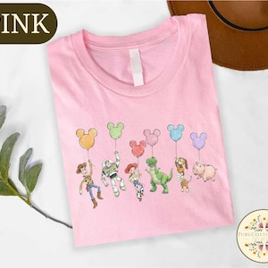 A pink t-shirt with a cartoon character design featuring a group of six Disney characters, including Woody, Buzz Lightyear, and other iconic figures, along with balloons.