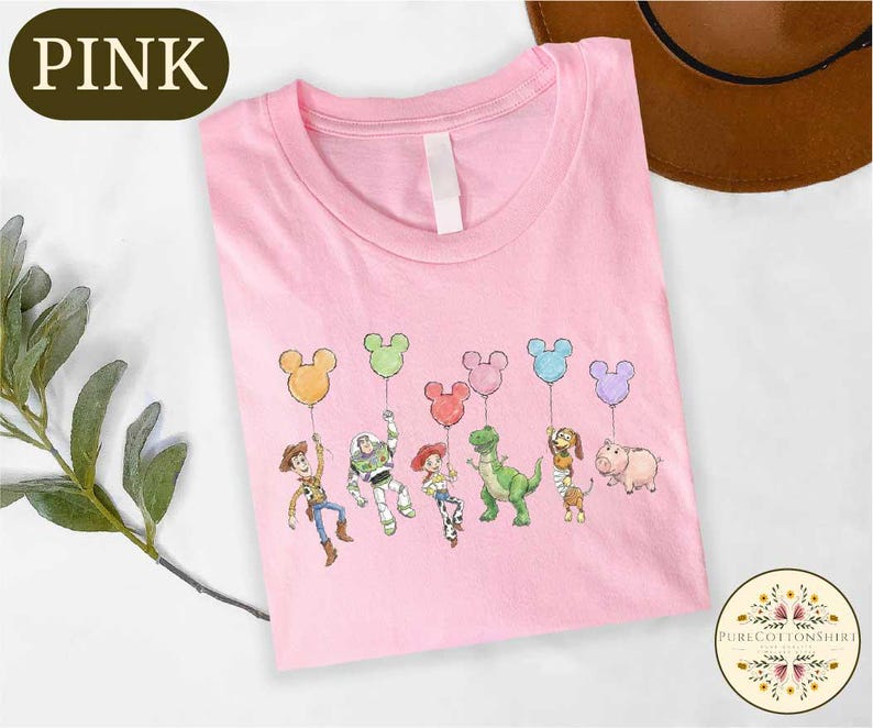 A pink t-shirt with a cartoon character design featuring a group of six Disney characters, including Woody, Buzz Lightyear, and other iconic figures, along with balloons.