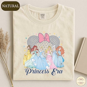 May include: Cream-colored t-shirt featuring a graphic of Disney princesses, a pink bow, and two sphere-shaped Mickey Mouse ears. The text "Princess Era" is printed in navy blue. The shirt is labeled "Natural" and "Comfort Colors".