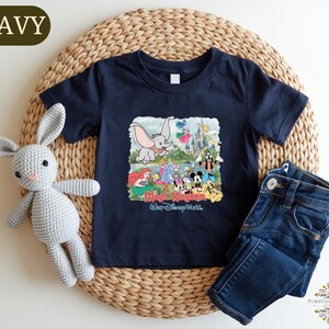 A navy blue t-shirt with a Disney World-themed graphic, a crocheted gray rabbit, and a pair of blue jeans are arranged on a woven placemat.