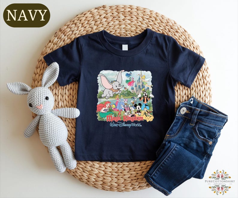 A navy blue t-shirt with a Disney World-themed graphic, a crocheted gray rabbit, and a pair of blue jeans are arranged on a woven placemat.