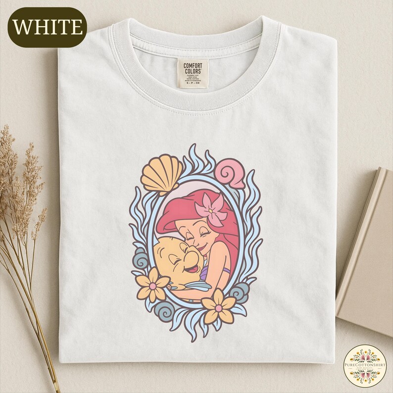 A white t-shirt with a colorful graphic design featuring a mermaid and a sea creature, surrounded by a decorative frame.