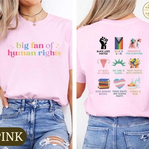 May include: Pink t-shirt with the text "big fan of human rights" in rainbow colors on the front. The back of the shirt features various graphic icons and phrases, including "Black Lives Matter" and "Love is Love".
