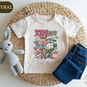 A white t-shirt with a colorful cartoon character design, a crocheted gray bunny, and a pair of blue jeans are arranged on a woven placemat.