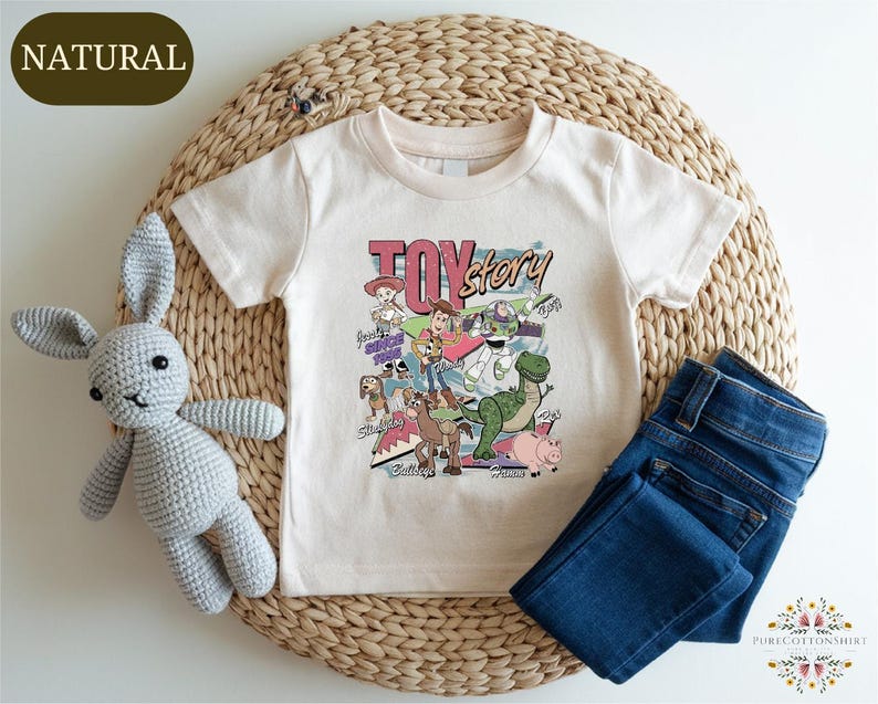A white t-shirt with a colorful cartoon character design, a crocheted gray bunny, and a pair of blue jeans are arranged on a woven placemat.