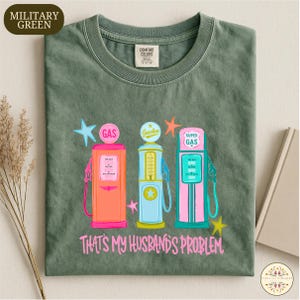 Pode incluir: T-shirt verde militar com um gráfico de três bombas de gasolina vintage coloridas. As bombas são rosa, azul e turquesa, com detalhes de estrelas e o texto "THAT'S MY HUSBAND'S PROBLEM" em rosa.