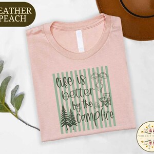 a pink t-shirt with a graphic design that says "life is better by the campfire" and includes a camping-related image. The t-shirt is laid out on a white surface, with a brown hat and some greenery visible in the background.