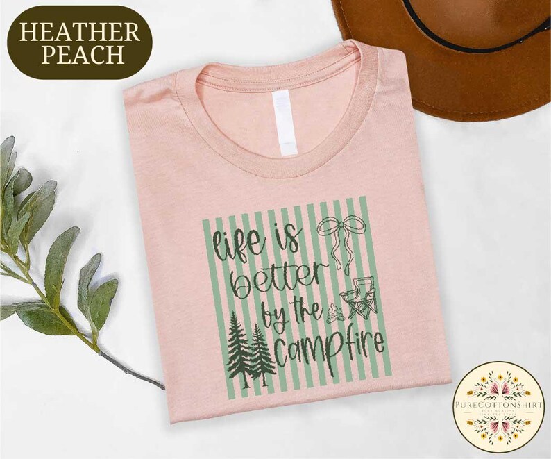 a pink t-shirt with a graphic design that says "life is better by the campfire" and includes a camping-related image. The t-shirt is laid out on a white surface, with a brown hat and some greenery visible in the background.