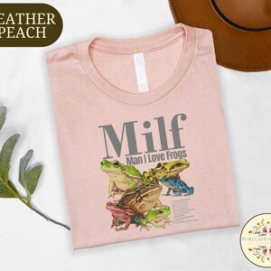 a pink t-shirt with a graphic of a frog and the text "Milf Man I Love Frogs" printed on it. The t-shirt is laid out on a white surface, with a brown hat and some greenery visible in the background.
