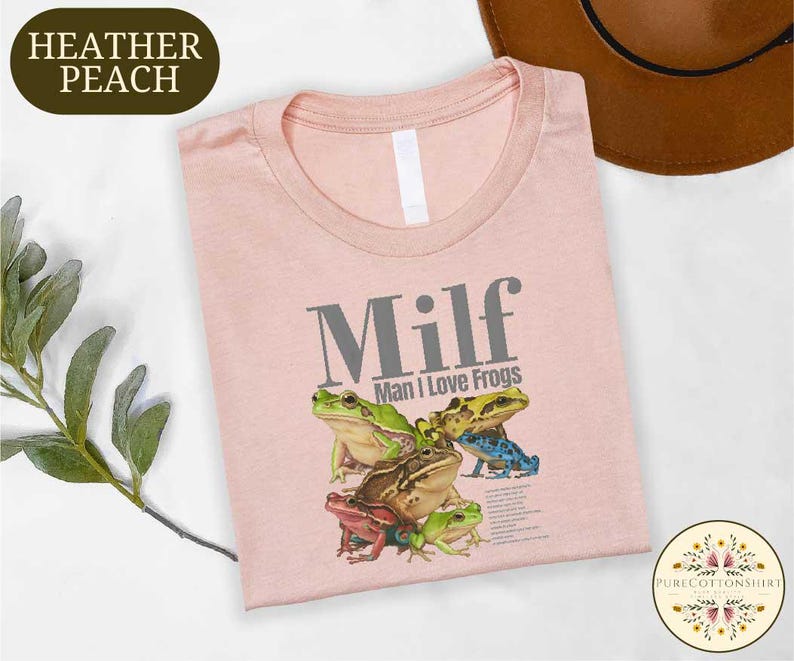a pink t-shirt with a graphic of a frog and the text "Milf Man I Love Frogs" printed on it. The t-shirt is laid out on a white surface, with a brown hat and some greenery visible in the background.