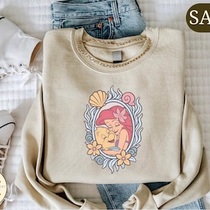 a casual outfit consisting of a beige sweater with a mermaid design, a pair of blue jeans, and white sneakers.