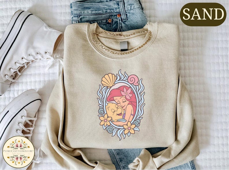 a casual outfit consisting of a beige sweater with a mermaid design, a pair of blue jeans, and white sneakers.
