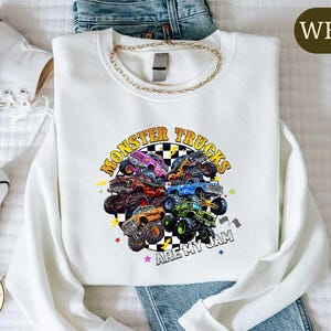 a white long-sleeved shirt with a graphic design of monster trucks, along with a pair of jeans and white sneakers.