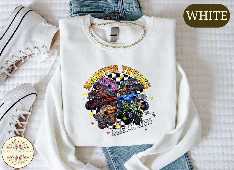 a white long-sleeved shirt with a graphic design of monster trucks, along with a pair of jeans and white sneakers.