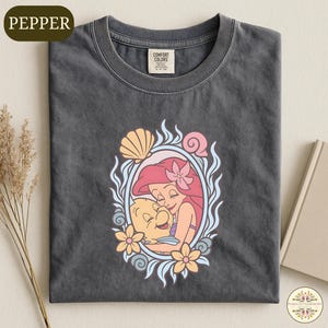 A gray t-shirt with a colorful graphic design featuring a mermaid and a sea creature, surrounded by flowers and a shell.