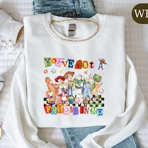 a white t-shirt with a colorful graphic design of characters from the Toy Story movie, along with the text "You've got a Fucked in Me" printed on it. The t-shirt is laid out on a bed, and there are some jeans and white sneakers visible in the background.