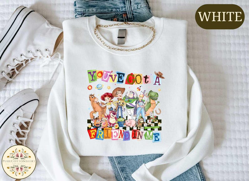 a white t-shirt with a colorful graphic design of characters from the Toy Story movie, along with the text "You've got a Fucked in Me" printed on it. The t-shirt is laid out on a bed, and there are some jeans and white sneakers visible in the background.