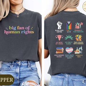 May include: Dark gray t-shirt with the text "big fan of human rights" in colorful letters. The back of the shirt features various social justice symbols and phrases, including "Black Lives Matter" and "Love is Love".