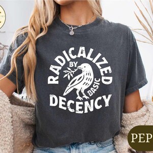 May include: Dark gray t-shirt with the text "RADICALIZED BY BASIC DECENCY" in a circular design around a dove holding an olive branch. The shirt is a pepper color.