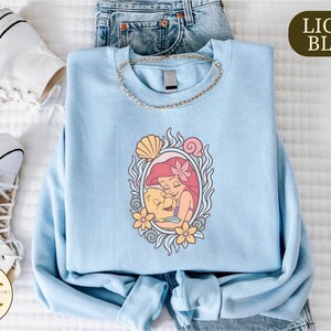 a light blue t-shirt with a mermaid design, a pair of white sneakers, and a pair of blue jeans, all laid out on a white surface.