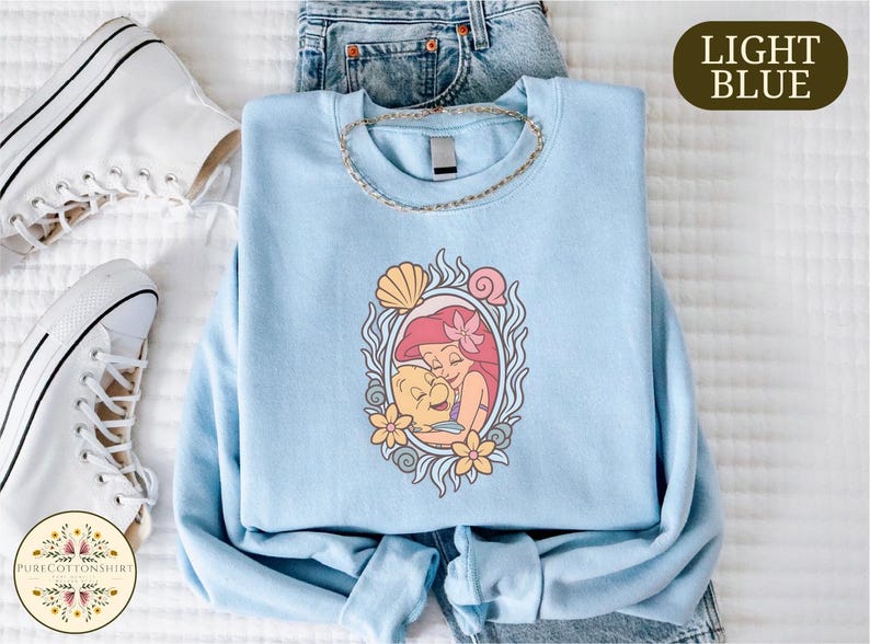 a light blue t-shirt with a mermaid design, a pair of white sneakers, and a pair of blue jeans, all laid out on a white surface.