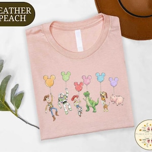 A pink t-shirt with a cartoon character design featuring a group of five Disney characters holding balloons, including a dinosaur, a pig, a piggy, a boy, and a girl.