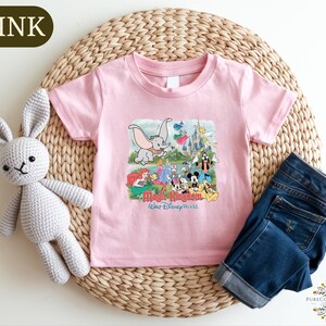 A pink t-shirt with a Disney-themed graphic, a crocheted white rabbit, and a pair of blue jeans are arranged on a woven placemat.