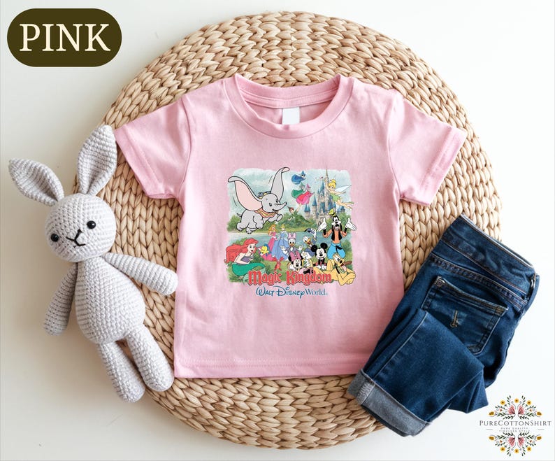 A pink t-shirt with a Disney-themed graphic, a crocheted white rabbit, and a pair of blue jeans are arranged on a woven placemat.