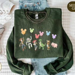 a green sweatshirt with a cartoon character design, including a character holding a balloon, a character holding a pig, and a character holding a dinosaur. The sweatshirt is placed on a bed, with a pair of white sneakers and a pair of blue jeans visible in the background.
