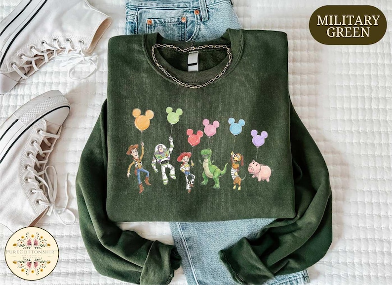 a green sweatshirt with a cartoon character design, including a character holding a balloon, a character holding a pig, and a character holding a dinosaur. The sweatshirt is placed on a bed, with a pair of white sneakers and a pair of blue jeans visible in the background.