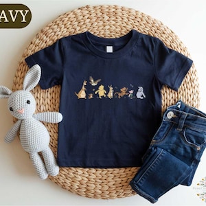 A navy blue t-shirt with a line of cartoon animal characters printed on it, a crocheted gray rabbit, and a pair of blue jeans are arranged on a woven placemat.