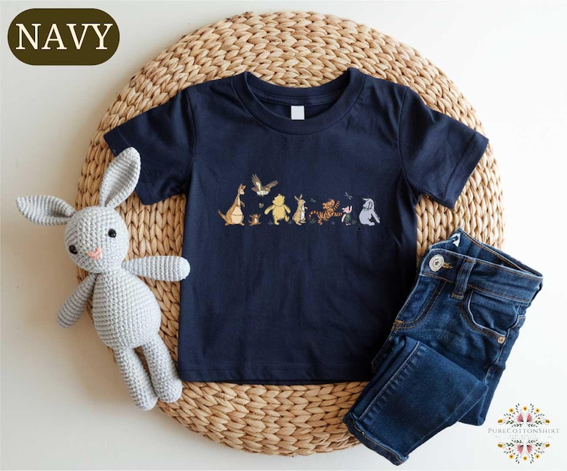 A navy blue t-shirt with a line of cartoon animal characters printed on it, a crocheted gray rabbit, and a pair of blue jeans are arranged on a woven placemat.