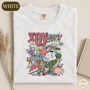 A white t-shirt with a colorful graphic design featuring characters from the Toy Story movie, including Woody, Buzz Lightyear, and other beloved characters.