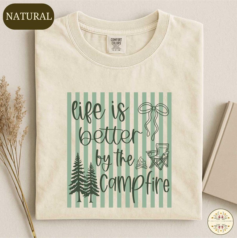 a beige t-shirt with a graphic design that says "life is better by the campfire" and includes a pine tree, a campfire, and a ribbon.