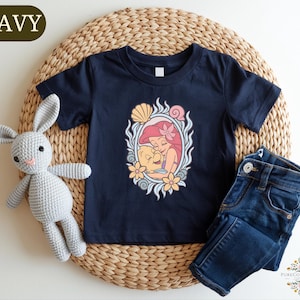 A navy blue t-shirt with a mermaid design, a gray crocheted bunny, and a pair of blue jeans are arranged on a woven placemat.