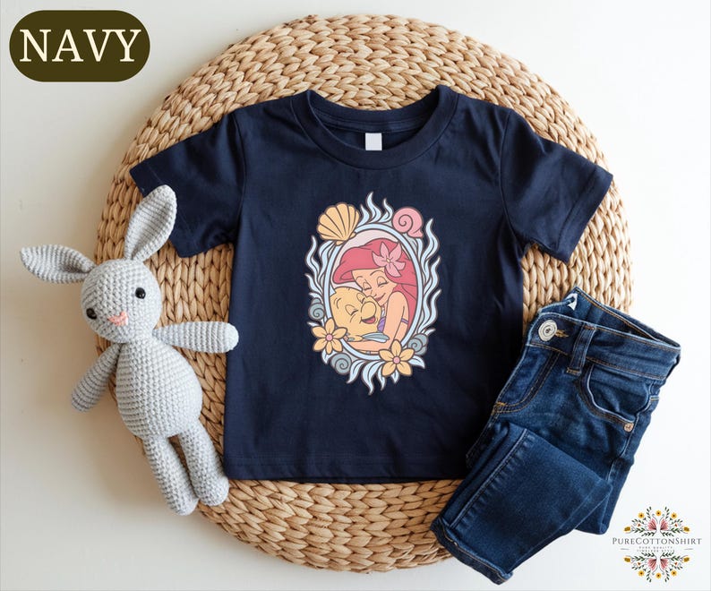 A navy blue t-shirt with a mermaid design, a gray crocheted bunny, and a pair of blue jeans are arranged on a woven placemat.