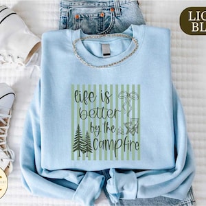 a light blue t-shirt with a graphic design, a pair of jeans, and a pair of white sneakers, all laid out on a bed.