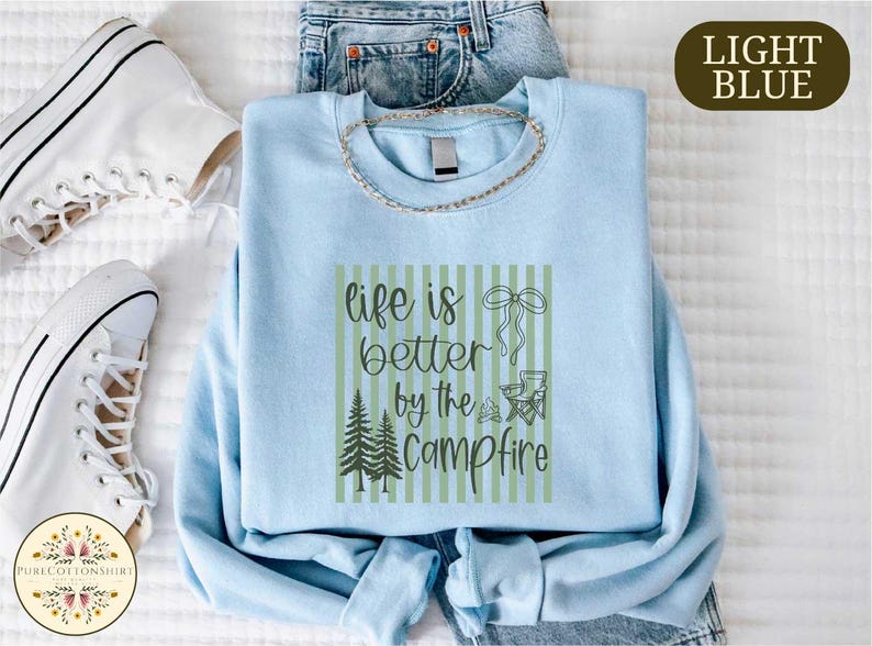 a light blue t-shirt with a graphic design, a pair of jeans, and a pair of white sneakers, all laid out on a bed.