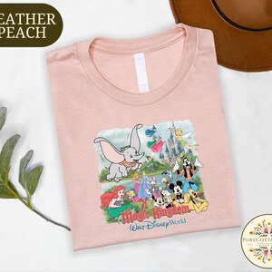 a pink t-shirt with a Disney-themed graphic, including characters from the Disney film "Magic Kingdom". The t-shirt is placed on a white surface, with a brown hat and some greenery visible in the background.