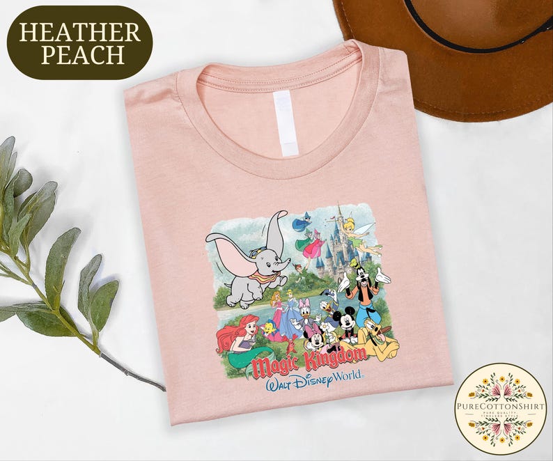 a pink t-shirt with a Disney-themed graphic, including characters from the Disney film "Magic Kingdom". The t-shirt is placed on a white surface, with a brown hat and some greenery visible in the background.