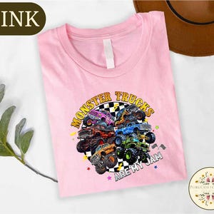 A pink t-shirt with a graphic design of monster trucks and the text "MONSTER TRUCKS ARE MY JAM" printed on it, along with a brown hat and some greenery in the background.