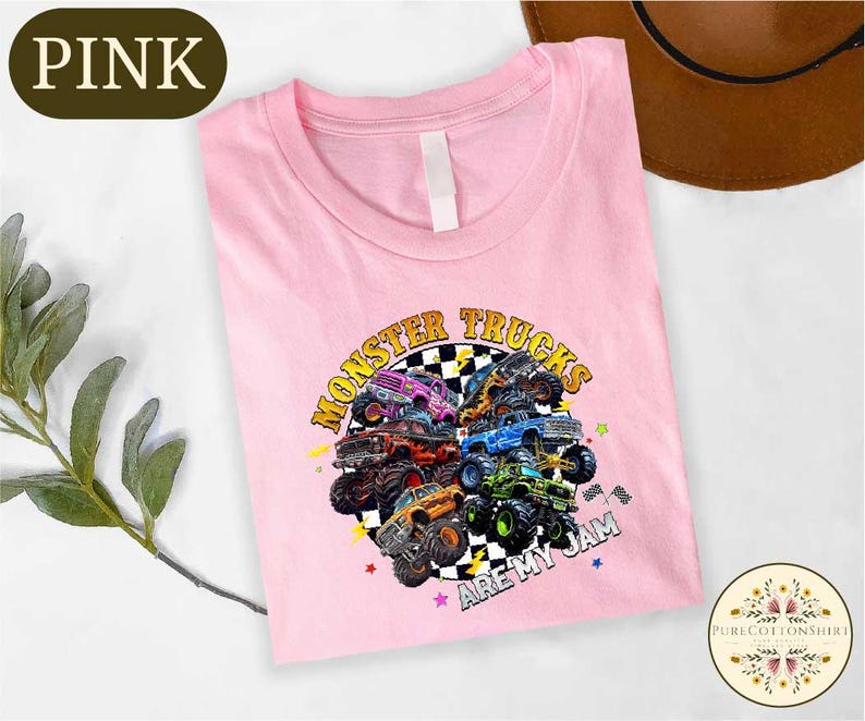 A pink t-shirt with a graphic design of monster trucks and the text "MONSTER TRUCKS ARE MY JAM" printed on it, along with a brown hat and some greenery in the background.