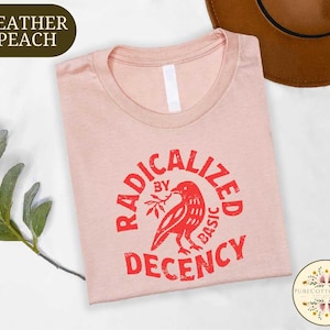 May include: A heather peach t-shirt with a crew neck, featuring a red graphic of a bird holding a branch, encircled by the words "RADICALIZED BY BASIC DECENCY." The shirt is folded and displayed on a white surface.